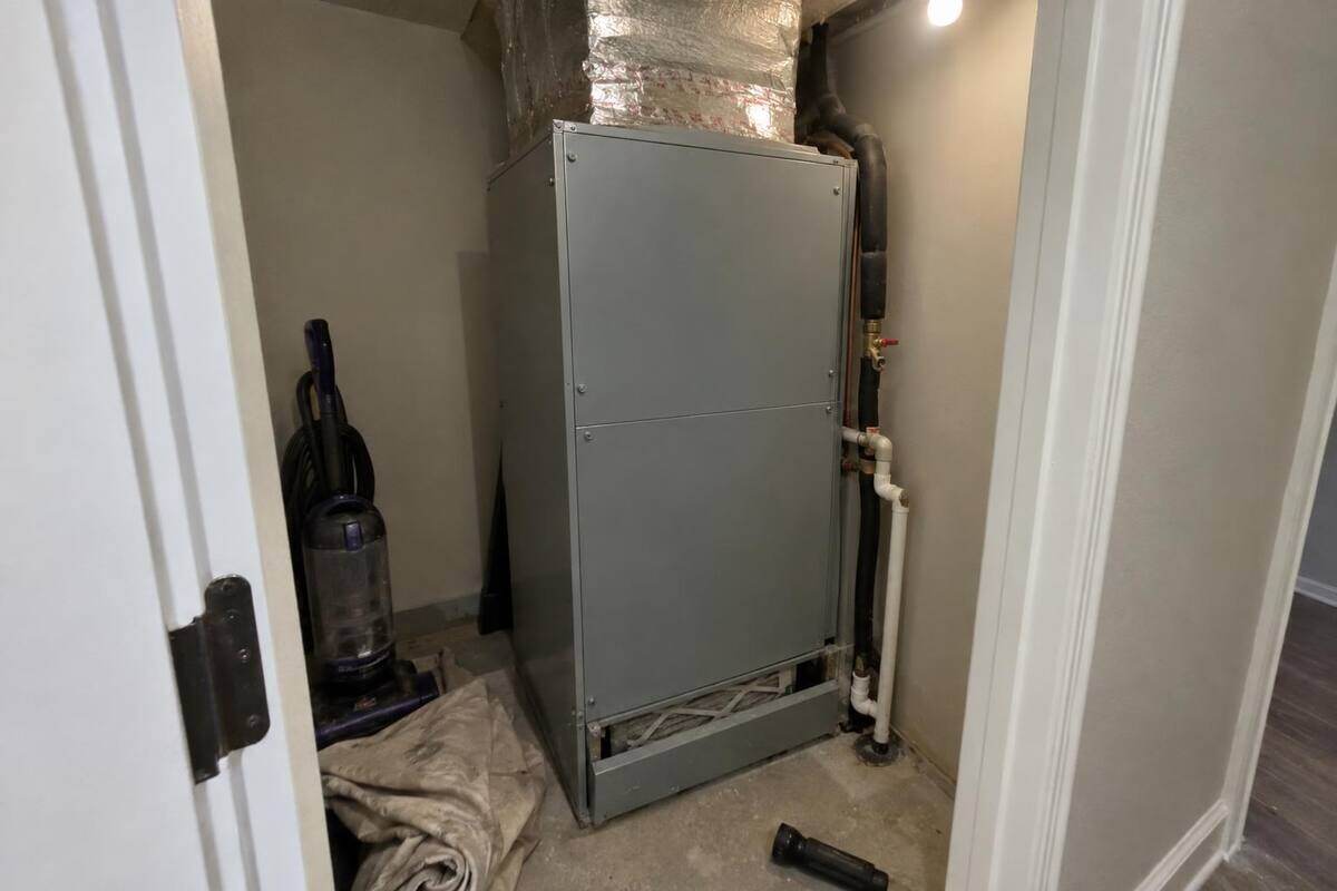 A closed gray air handler cabinet standing in a narrow hall closet with a dusty filter edge showing at the bottom