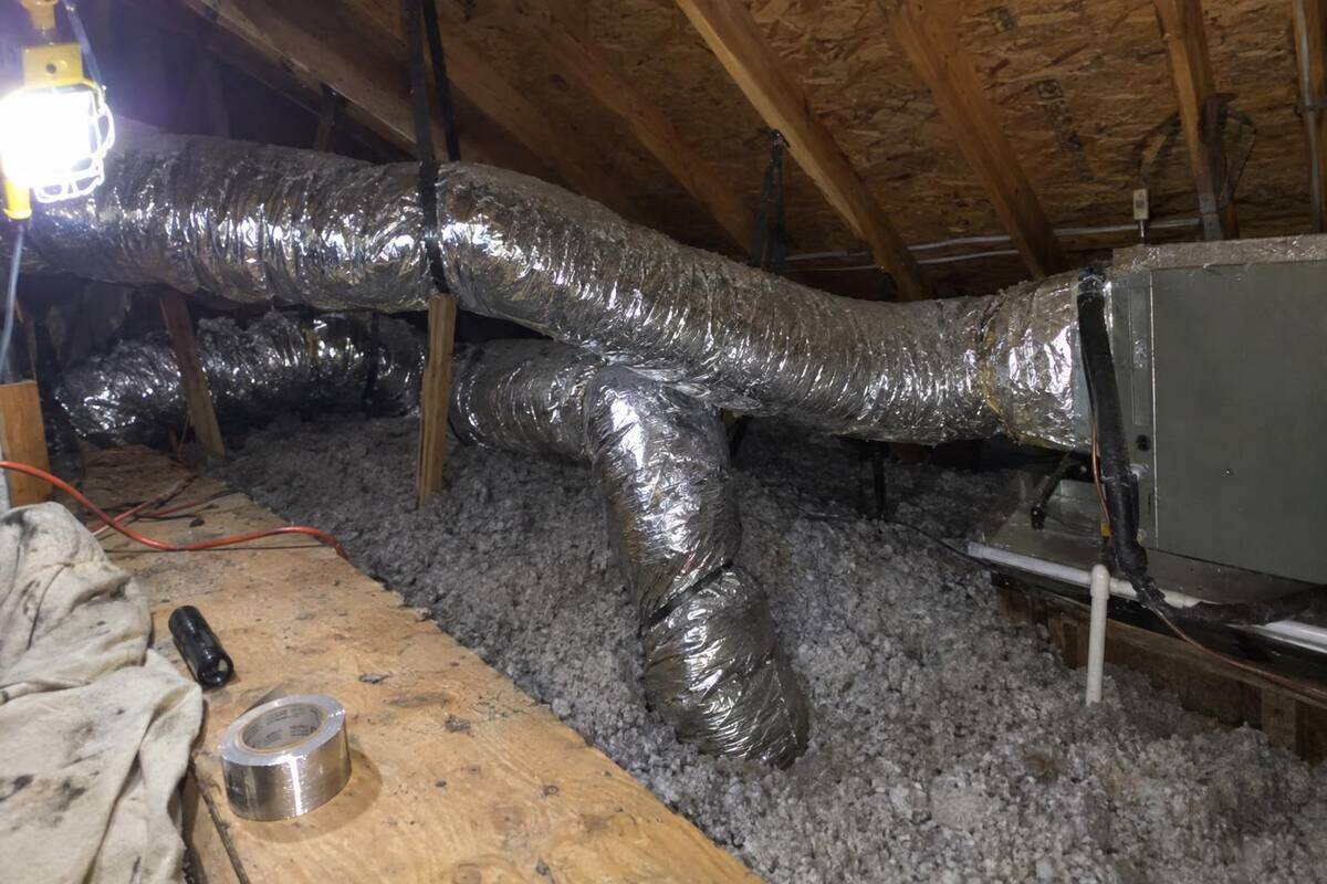 Silver flex duct runs sagging between rafters over blown-in insulation in an attic lit by a clip-on work light