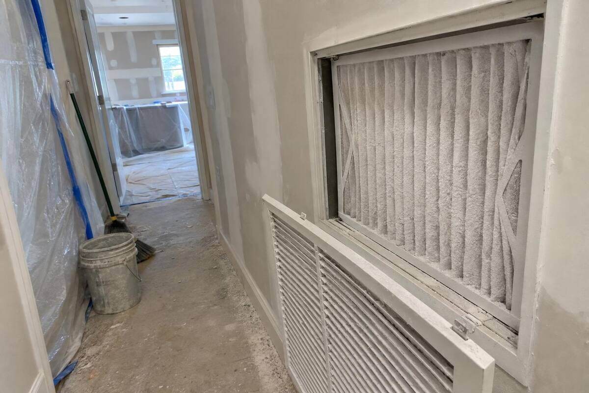 A hallway return grille swung open with its filter coated white in drywall dust and a plastic-sheeted remodel beyond
