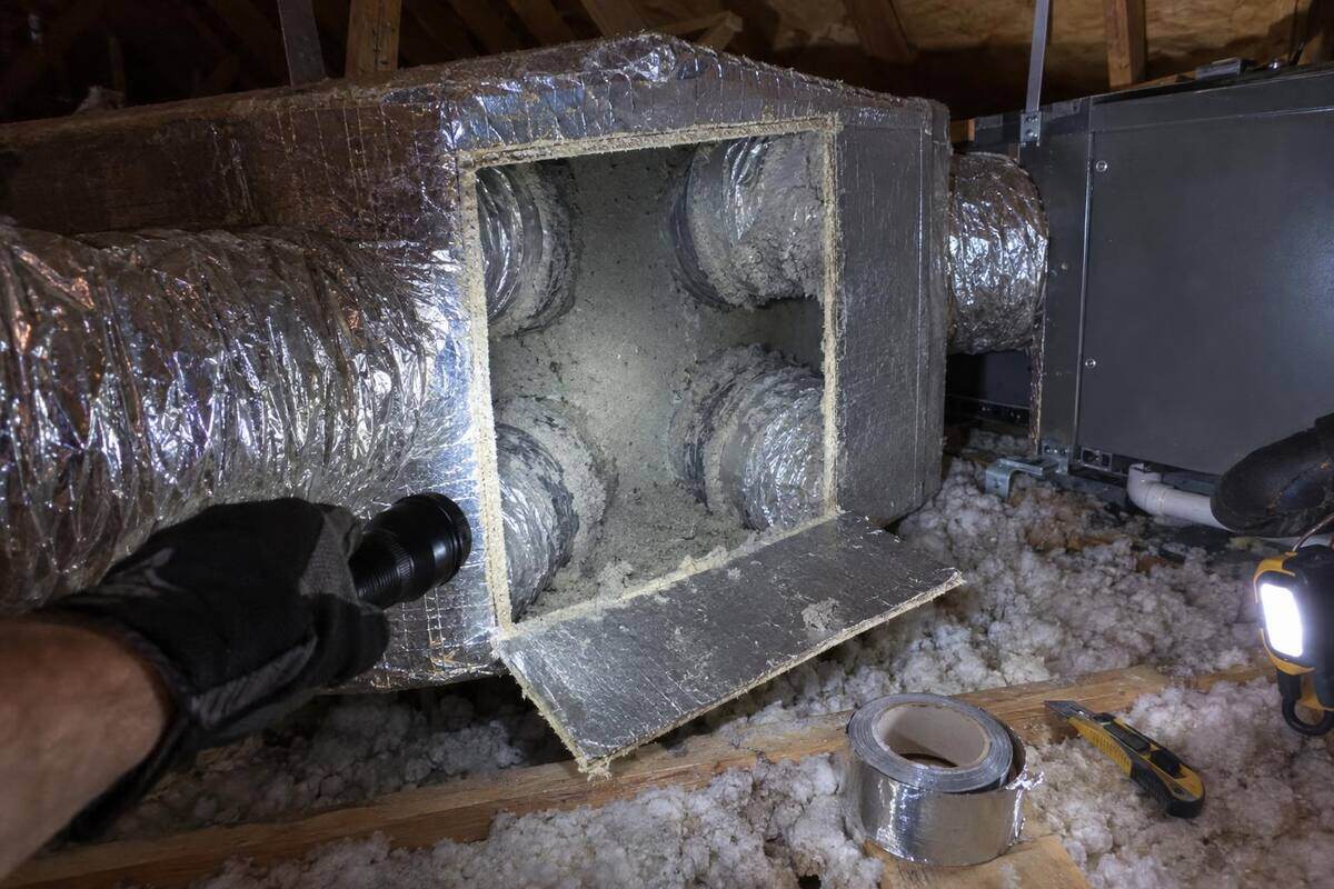 A ductboard plenum box in an attic with an access panel folded open and a flashlight lighting the dusty collars inside